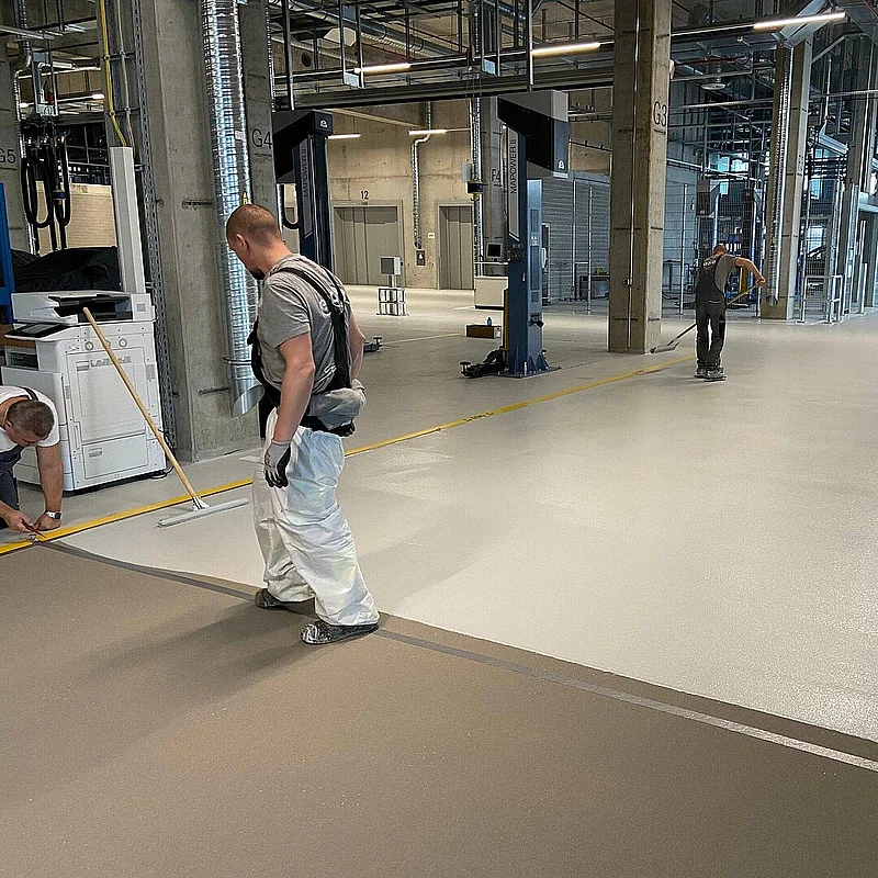 Arbeiter versiegelt eine beschichtete Bodenfläche in einer Industriehalle als abschließenden Arbeitsschritt.
